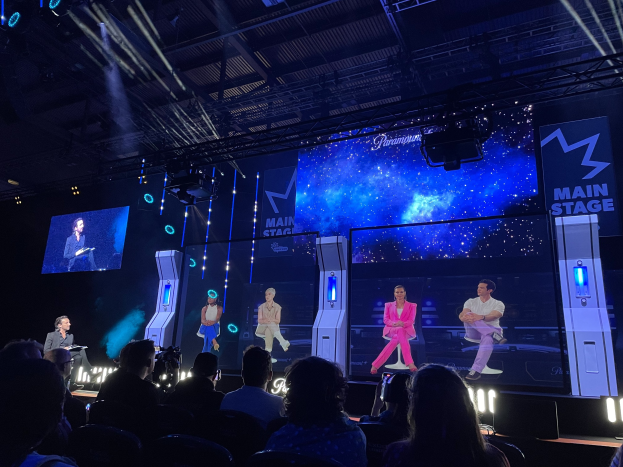 Eine Gruppe von Menschen auf einer hell erleuchteten Bühne mit Bildschirmen, einer Person mit Mikrofon und anderen in Stühlen, unter einer Decke mit Beleuchtungsausrüstung, was auf ein Gaming-Event hindeutet.