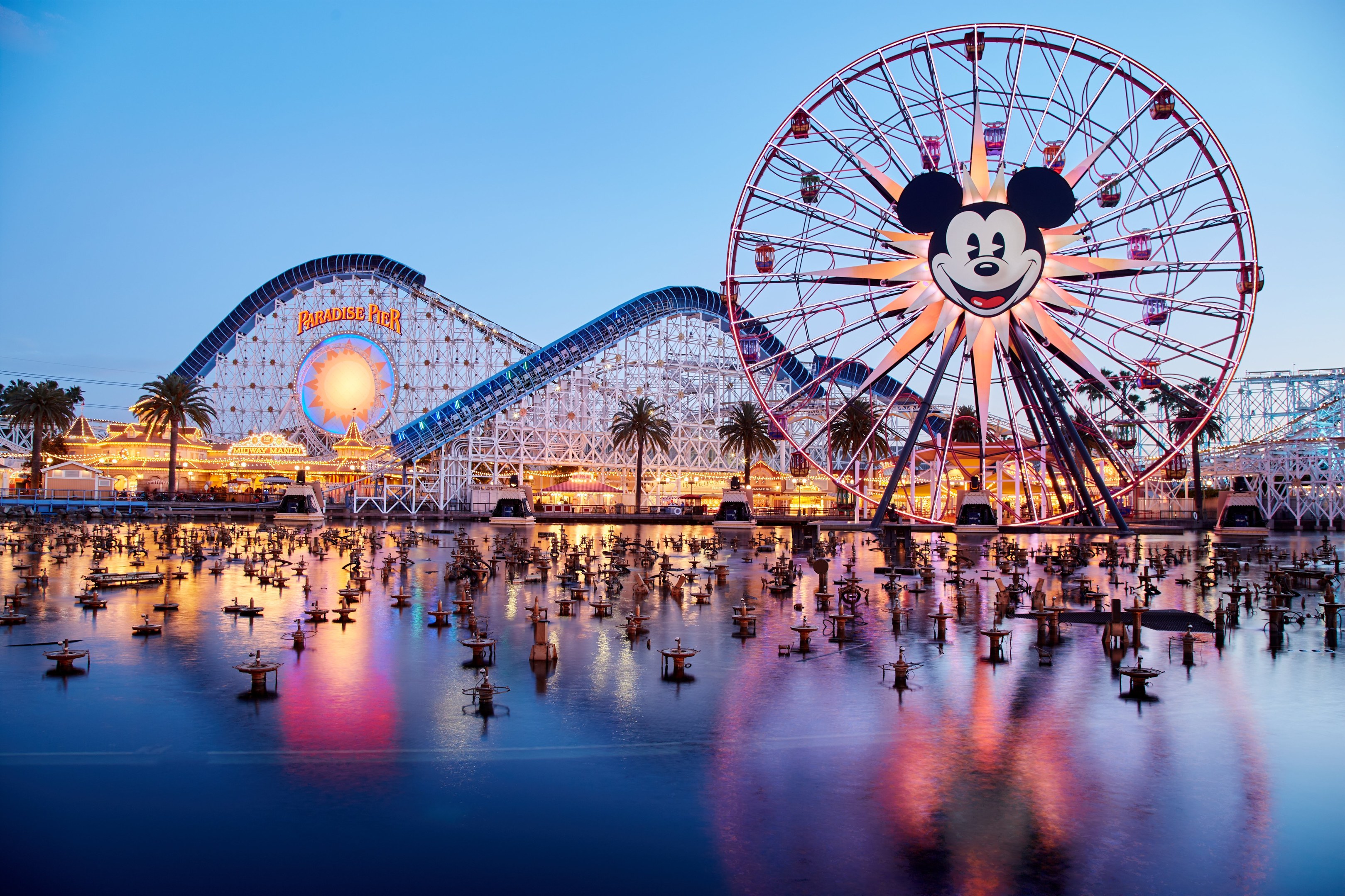 A vibrant dusk scene at Disneyland's California Adventure featuring a giant Mickey Mouse wheel in the foreground, a roller coaster in the background, and boats on the water, with a colorful sky and silhouetted trees.