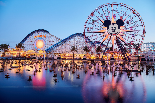 A vibrant dusk scene at Disneyland's California Adventure featuring a giant Mickey Mouse wheel in the foreground, a roller coaster in the background, and boats on the water, with a colorful sky and silhouetted trees.