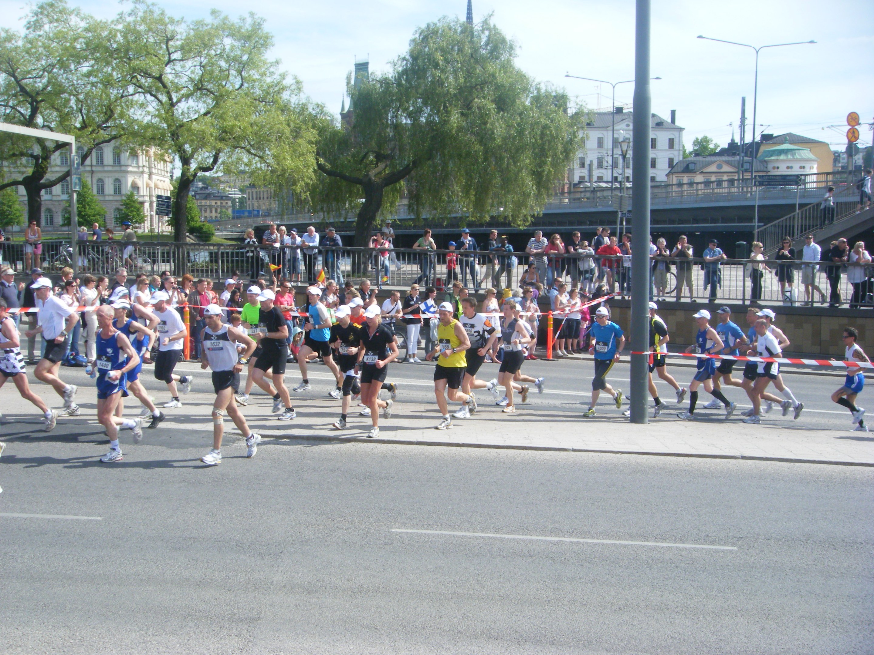 Group of people running in a marathon on a road with goal post, ribbon, barricades, poles, signboards, bridge, buildings, trees, and a cloudy sky.