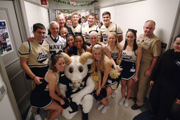 Gruppe von Menschen, die mit einem maskierten Maskottchen posieren, umgeben von Cheerleadern, Plakat an der linken Wand, Türen an der rechten Seite und Hintergrund-Elemente einschließlich einer Wand, einer Schautafel, Dekorationen und Deckenleuchten.