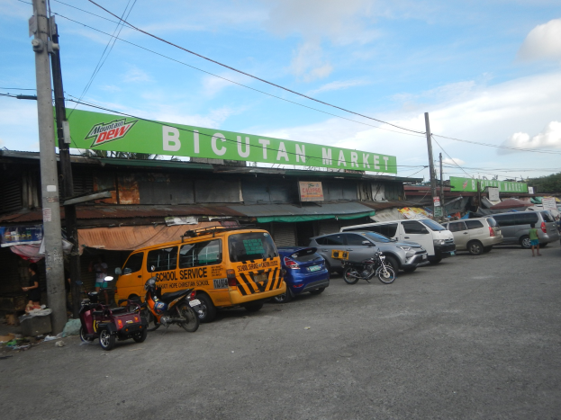 Eine belebte Straßenszene auf dem Bicutan Market in Cebu City, Philippinen, mit Fahrzeugen, Fußgängern, Strommasten, Gebäuden, Bäumen und einem bewölkten Himmel.