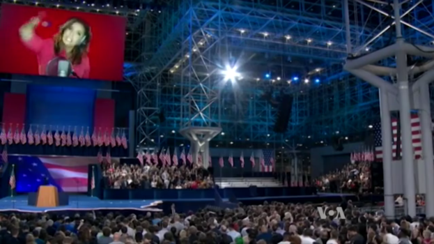 Eine große Menschenmenge sitzt vor einer Bühne mit einem Podium, einem Mikrofon, Flaggen, einem Bildschirm mit dem Bild einer Frau und Lichtausrüstung, wahrscheinlich bei einer Präsidenten-Debatte.