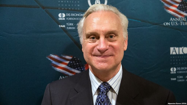 Ein Mann im Anzug und Krawatte steht lächelnd vor einer blauen Wand mit einem Banner im Hintergrund.