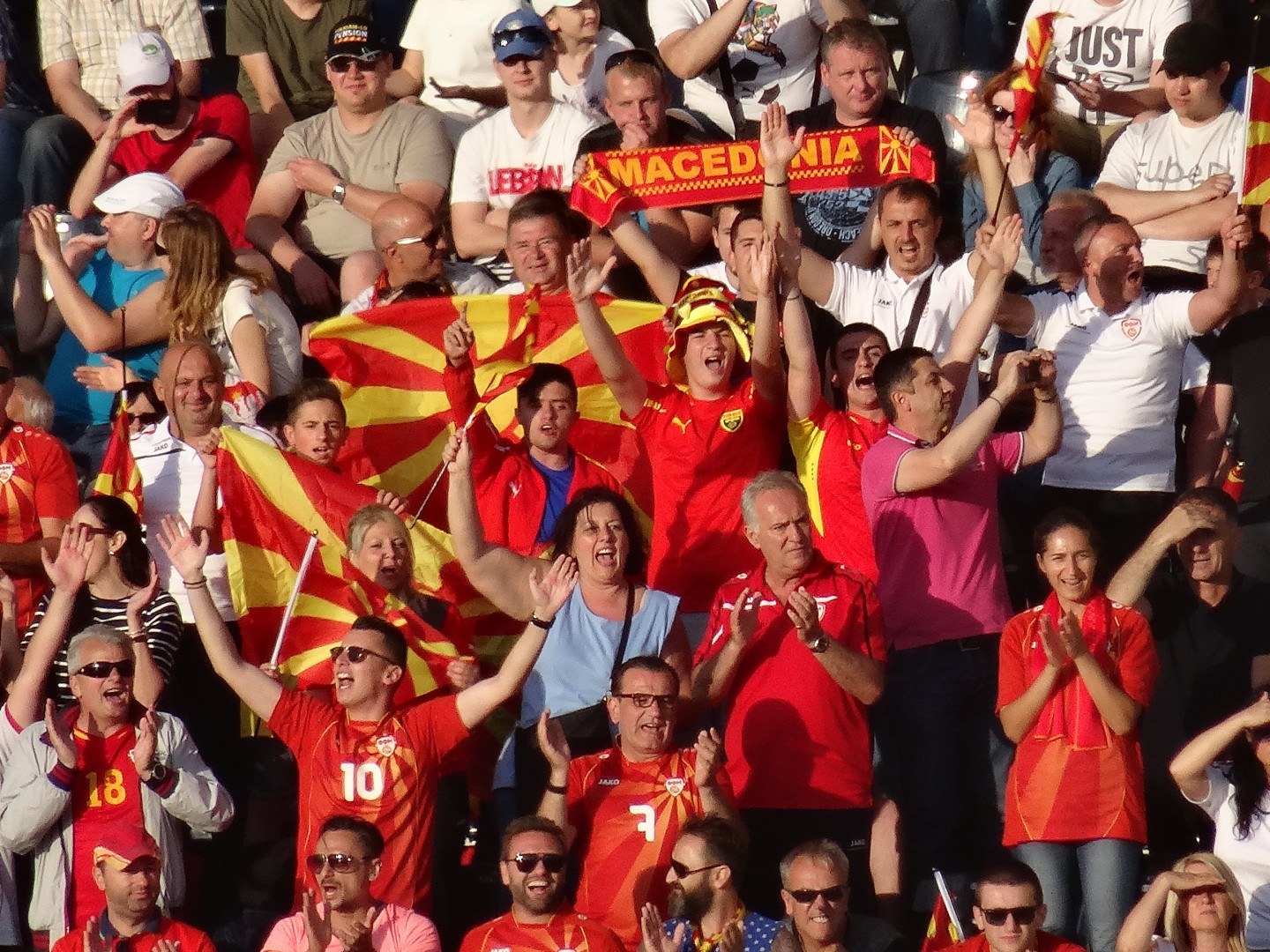 Eine Gruppe von Menschen in einem Stadion, die ihre Hände heben, einige tragen Kappen und Schutzbrillen und halten Fahnen und Schilder mit Text, im Hintergrund sind Geländer zu sehen.