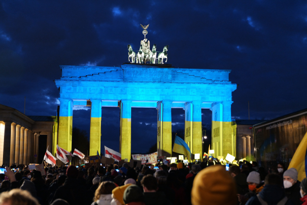 Menschenmenge mit Fahnen und Plakaten vor dem Brandenburger Tor in Berlin, mit einem Banner auf der rechten Seite.