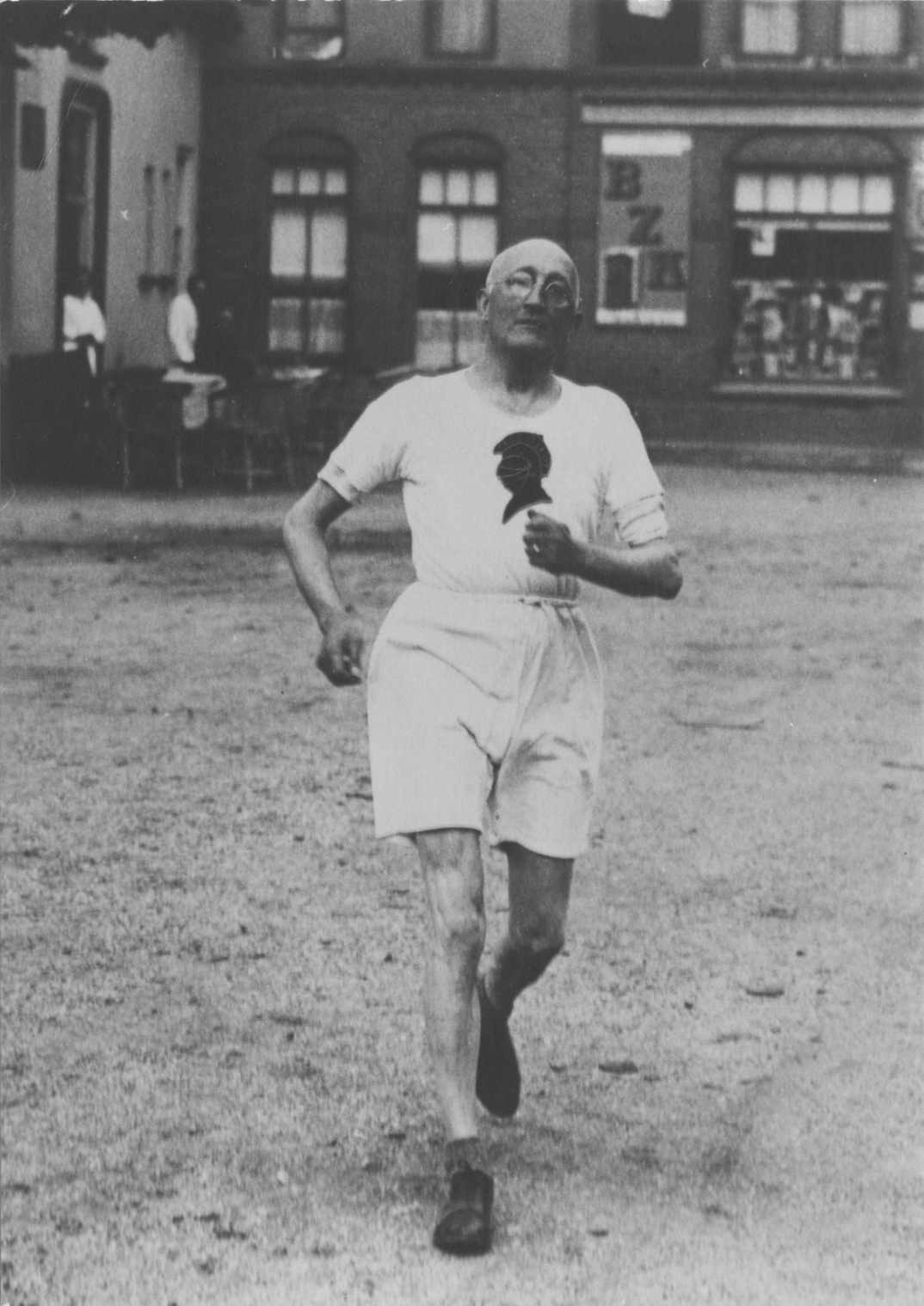 Schwarzes und weißes Foto eines Mannes, der bei einem Rennen läuft, mit einem Gebäude im Hintergrund und Zuschauern um einen Tisch herum, der angeblich der Gründer des Boston-Marathons ist.