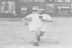 Schwarzes und weißes Foto eines Mannes, der bei einem Rennen läuft, mit einem Gebäude im Hintergrund und Zuschauern um einen Tisch herum, der angeblich der Gründer des Boston-Marathons ist.