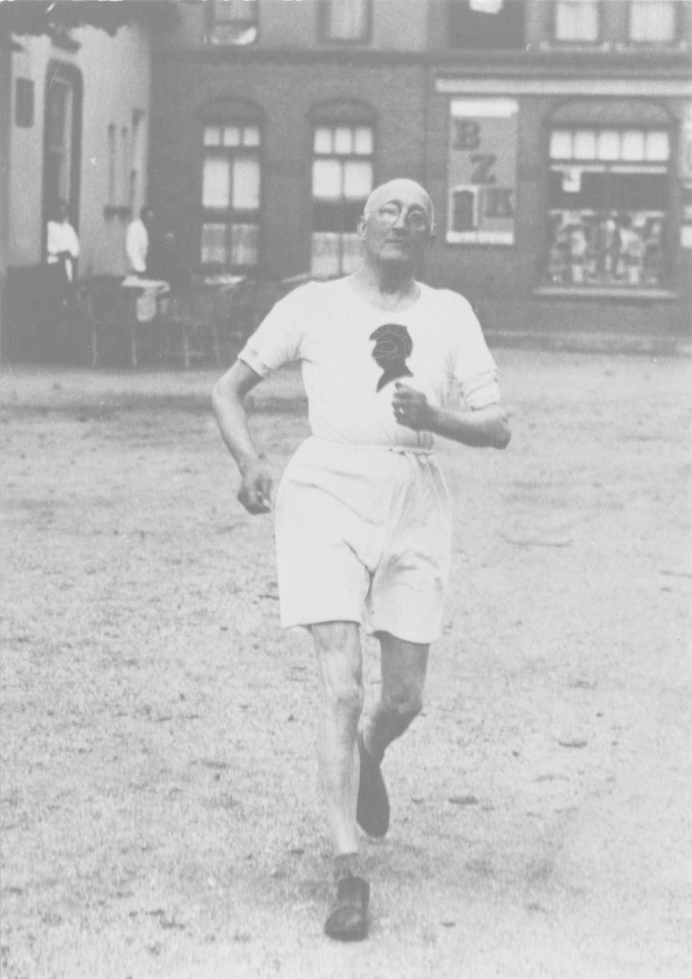 Schwarzes und weißes Foto eines Mannes, der bei einem Rennen läuft, mit einem Gebäude im Hintergrund und Zuschauern um einen Tisch herum, der angeblich der Gründer des Boston-Marathons ist.