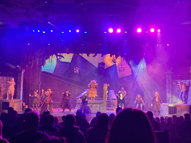 Eine Gruppe von Menschen in Kostümen auf einer hell erleuchteten Bühne über einer Sitzreihe mit einer Wand im Hintergrund.