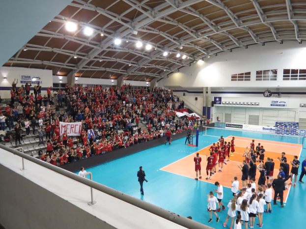 Eine Gruppe von Menschen spielt Volleyball in einer Turnhalle mit einem Netz, Zuschauern auf Stühlen, Bannern und einer Wand mit Fenstern und Deckenleuchten im Hintergrund.
