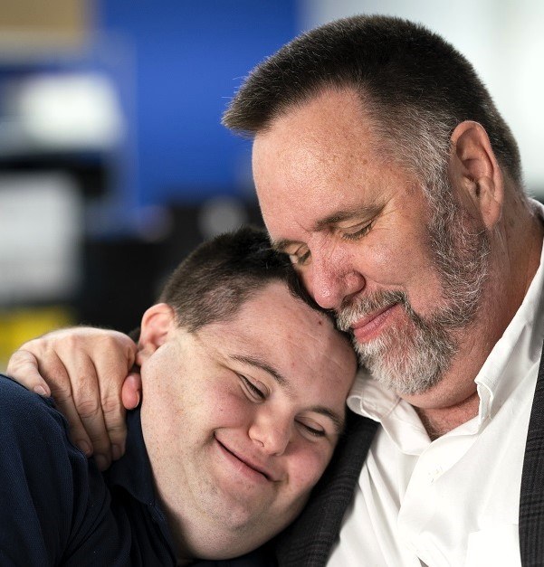 Zwei Personen, ein Mann in einem weißen Hemd und schwarzer Jacke und ein Junge, umarmen sich in einer herzlichen Umarmung mit einem unscharfen Hintergrund.