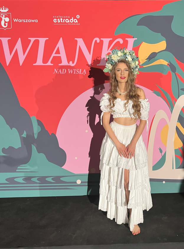 Eine Frau in einem weißen Kleid mit einer Blumenkrone steht auf einem roten Teppich, mit einer Tafel im Hintergrund, auf der "Wlanci 2019 - Ankünfte auf dem roten Teppich" steht.