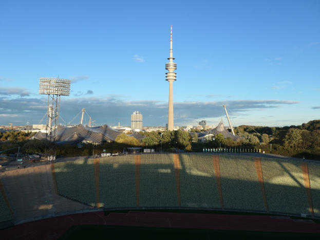 Luftaufnahme des Olympiastadions in Berlin mit dem Fernsehturm im Hintergrund, umgeben von Bäumen, Gebäuden und beleuchteten Bereichen unter einem bewölkten Himmel.