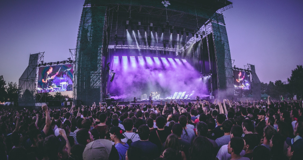 Eine große Menge Menschen bei einem Musikfestival schaut auf einer hell beleuchteten Bühne mit Instrumenten und Bildschirmen, die vor einem Gebäude, Bäumen und einem klaren blauen Himmel aufgebaut sind.