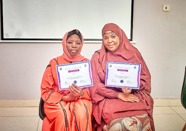 Zwei Frauen sitzen nebeneinander und lächeln, während sie Zertifikate halten; ein Stuhl ist auf der rechten Seite, ein wandmontiertes Objekt und ein Projektionsschirm sind im Hintergrund zu sehen, was auf eine Universität hindeutet, wo sie gerade Zertifikate von der Universität Nigeria erhalten haben.