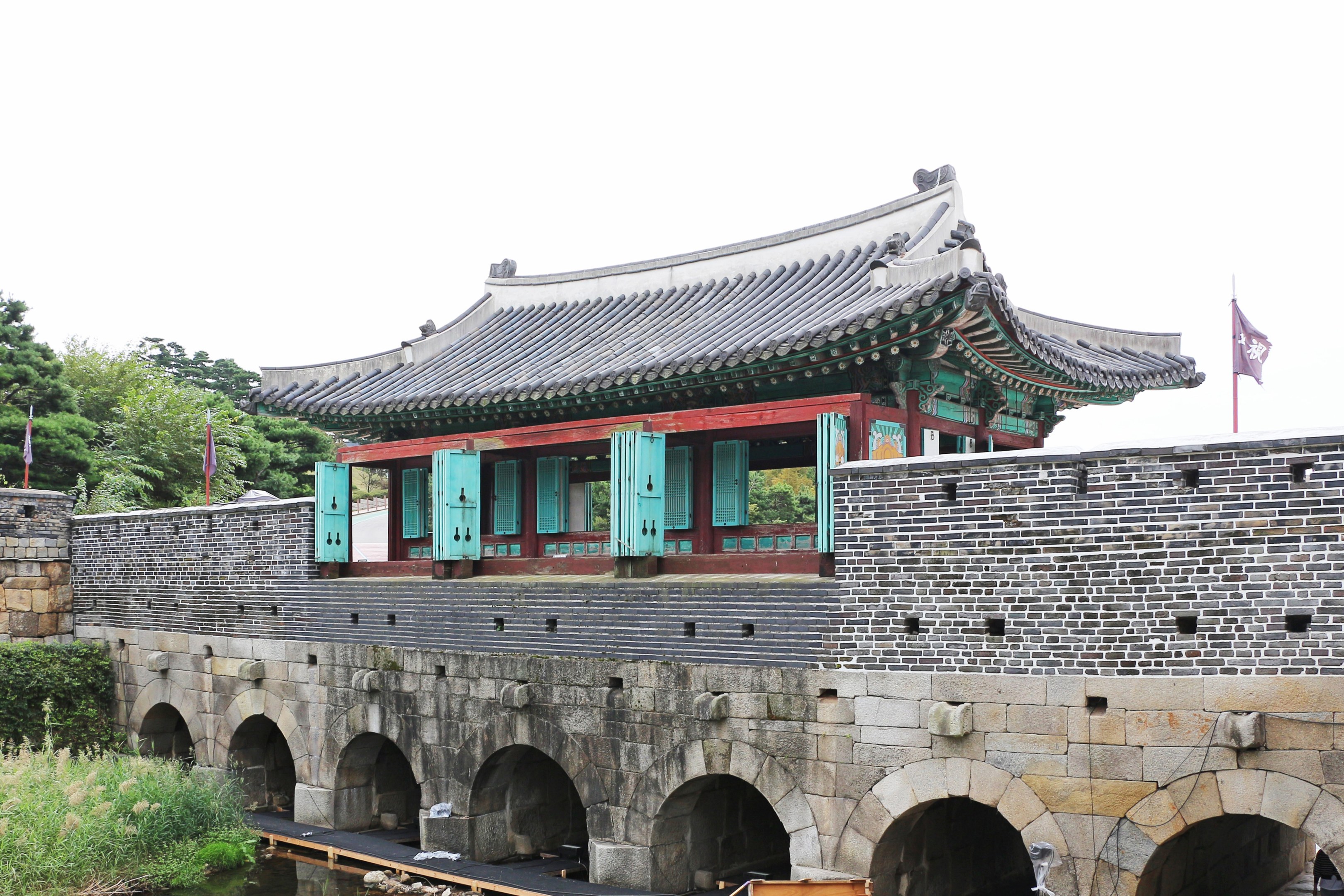 Ein traditioneller Palastkomplex in Seoul, Südkorea, mit einer Brücke über einen Teich, umgeben von Grünflächen und geschmückt mit Flaggen unter einem klaren Himmel.