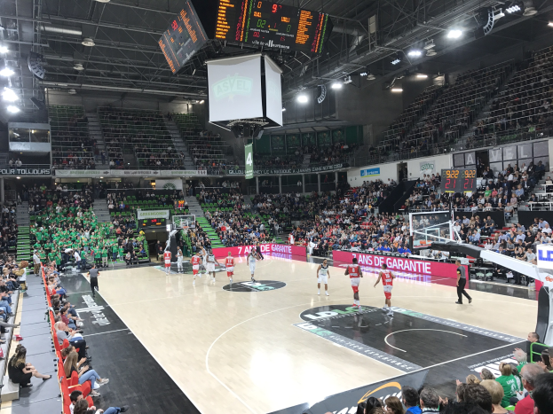 Ein Basketballspiel in einer großen Arena mit Zuschauern auf den Tribünen, Anzeigetafeln mit "Asvel vs Galatasaray" und einer beleuchteten Decke mit einem Bildschirm über dem Court.
