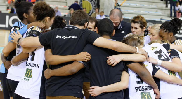 Eine Gruppe von Frauen-Basketballspielerinnen steht zusammen auf einem Platz, einige stehen, andere sitzen auf einer Treppe im Hintergrund und feiern einen Sieg.