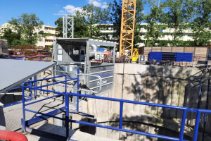 Eine Baustelle mit einem großen Metallgerüst, umgeben von einer Wand, einem Geländer, Rohren und anderen Gegenständen, mit Bäumen, Gebäuden mit Fenstern und einem bewölkten Himmel im Hintergrund, das die Baustelle der neuen Wasseraufbereitungsanlage im Disney's Hollywood Studios zeigt.