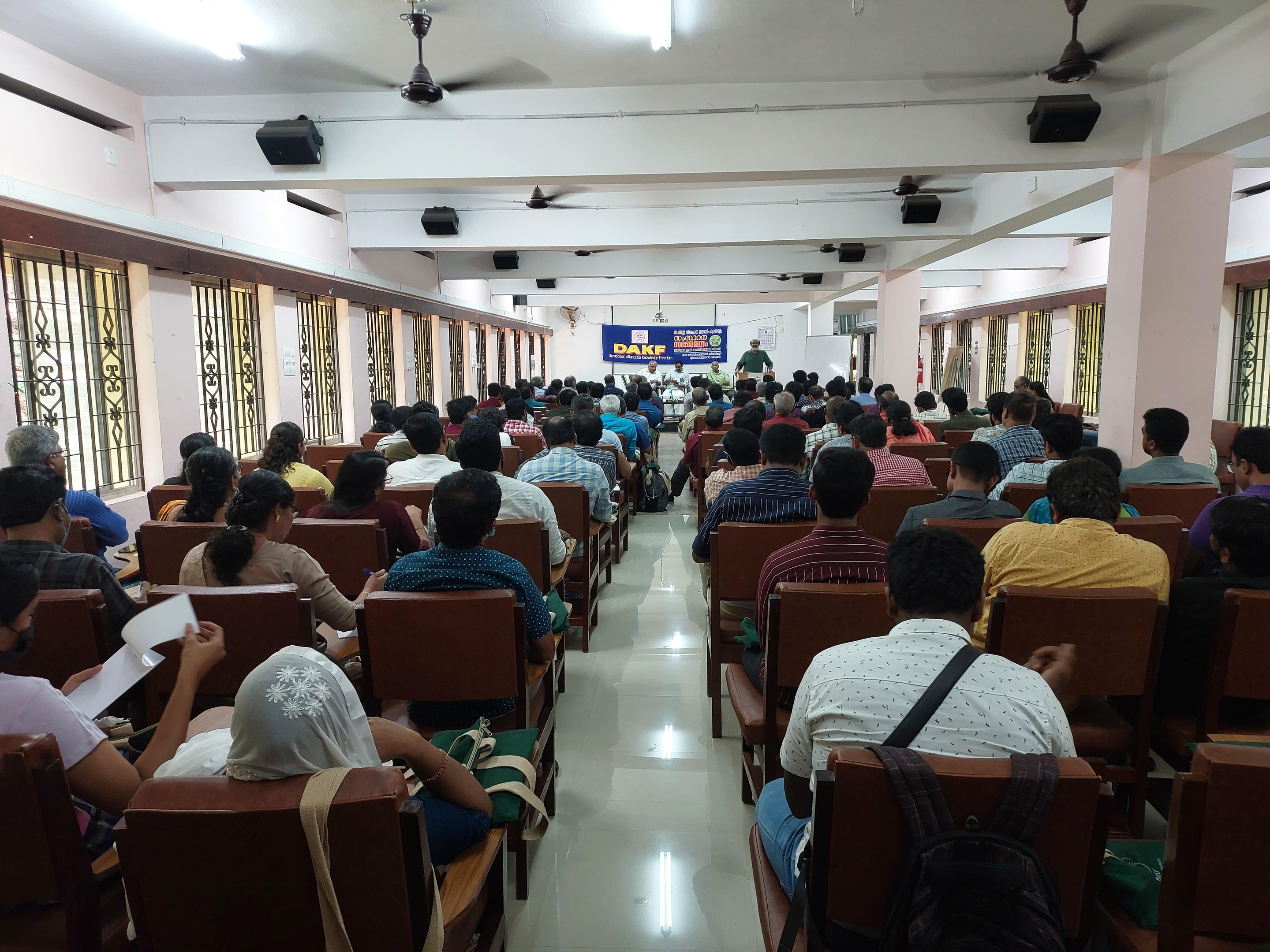 Eine große Gruppe von Menschen, die in einem Raum mit Fenstern auf der linken und rechten Seite, Säulen in der Rückseite und einer zentralen Fahne sowie Deckenventilatoren und -leuchten sitzen, was auf eine Workshop-Veranstaltung über Bildung in Indien hinweist.