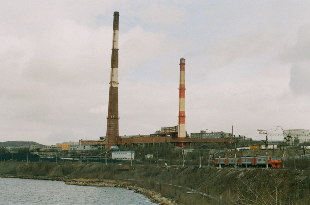 Steinkohlekraftwerk neben einem Gewässer mit einem Zug auf einem Bahngleis, Bäumen, Gebäuden, Strommasten und Übertragungsmasten im Vordergrund und einem bewölkten Himmel im Hintergrund.