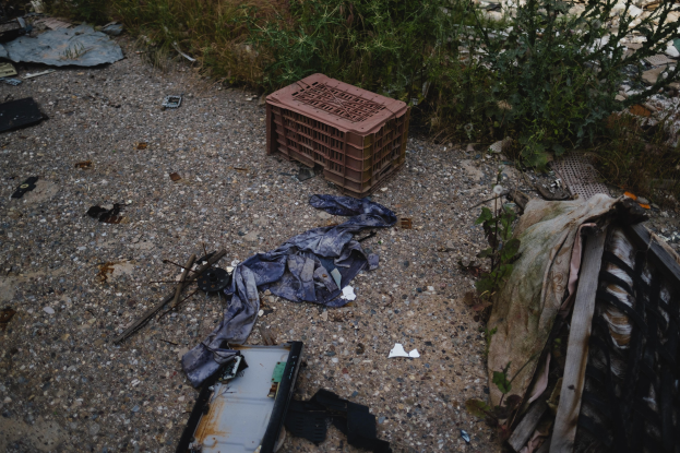 Ein Haufen Müll, einschließlich eines Plastikbehälters, Kleidung und anderen Gegenständen, der auf dem Boden neben einem Zaun verstreut ist, mit Pflanzen und Gras im Hintergrund.