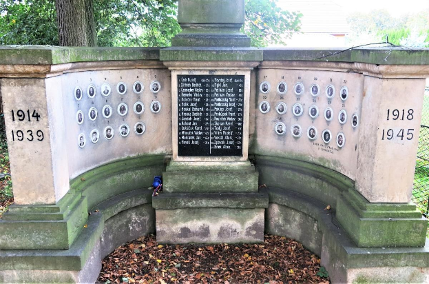 Ein Holocaust-Gedenkstein in einem jüdischen Friedhof in Berlin, mit einer Tafel mit Text und Zahlen, umgeben von Bäumen, einem Zaun und verstreuten trockenen Blättern.