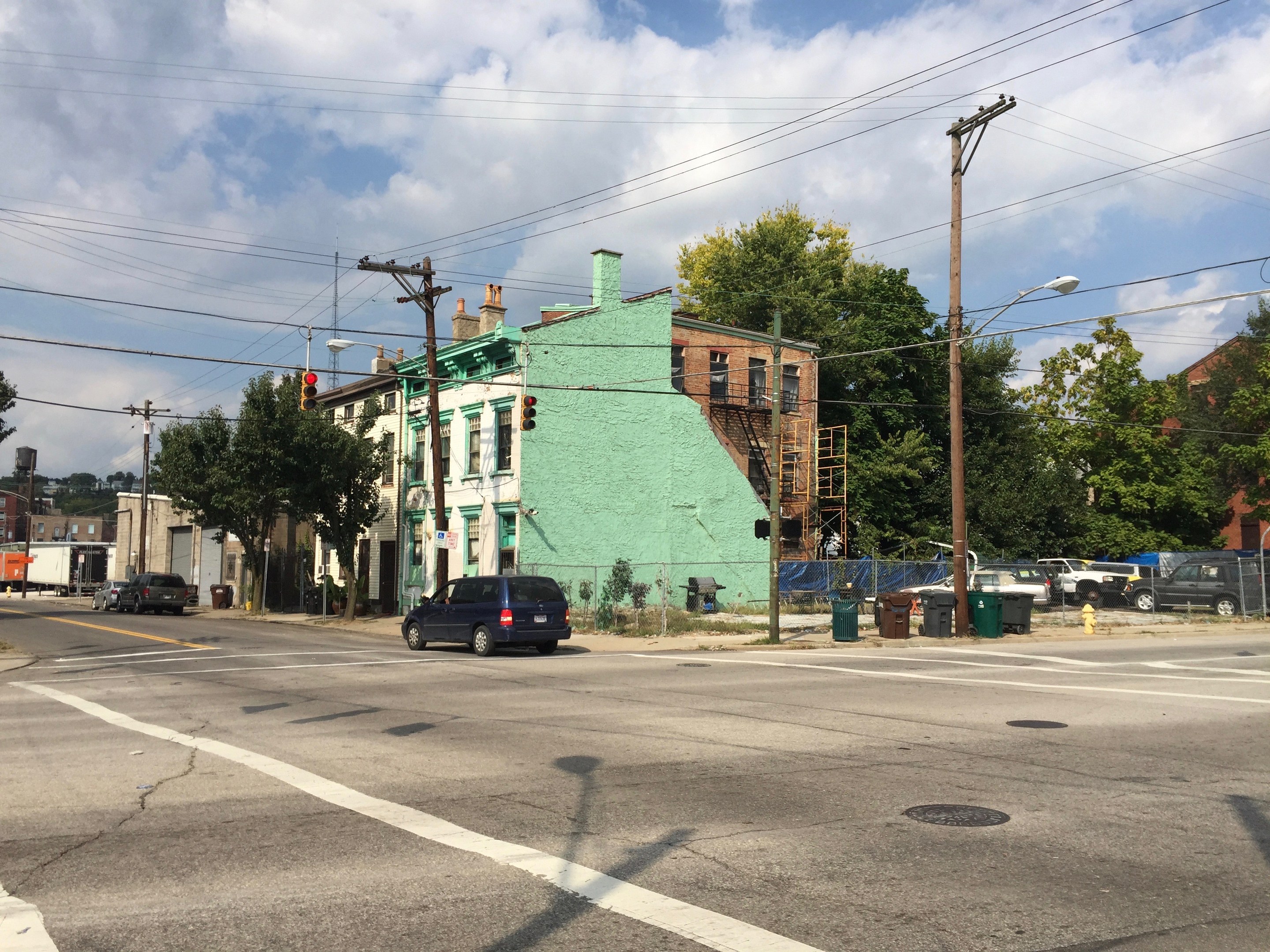 A street corner with parked cars, electric poles, light poles, traffic signals, dustbins, trees, buildings, and a cloudy sky, featuring a green apartment complex in the foreground.