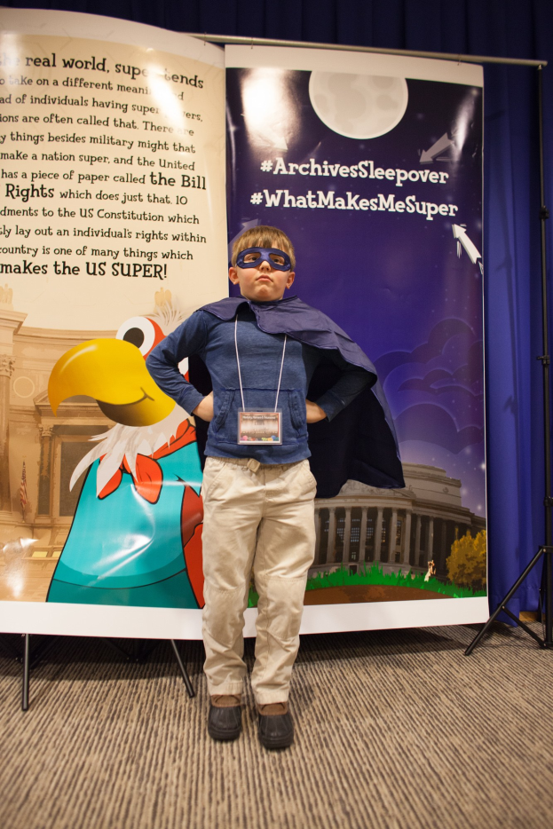 Junge in Superheldenkostüm vor einem Banner mit Text und einem Vogelbild, mit Schuhen und einem Vorhang im Hintergrund.