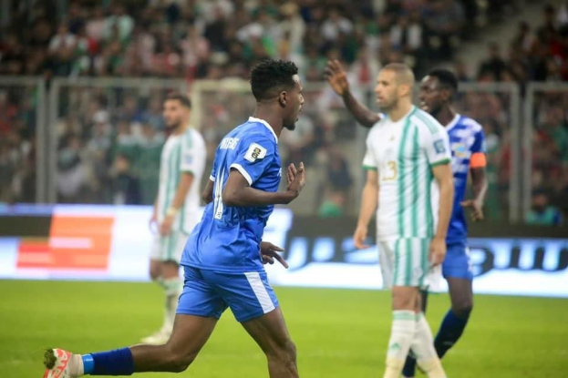 Ein Fußballspieler in einem blauen und weißen Trikot feiert ein Tor, während er von Teamkollegen in weißen und grünen Trikots umgeben ist, mit einer jubelnden Menge im Hintergrund.