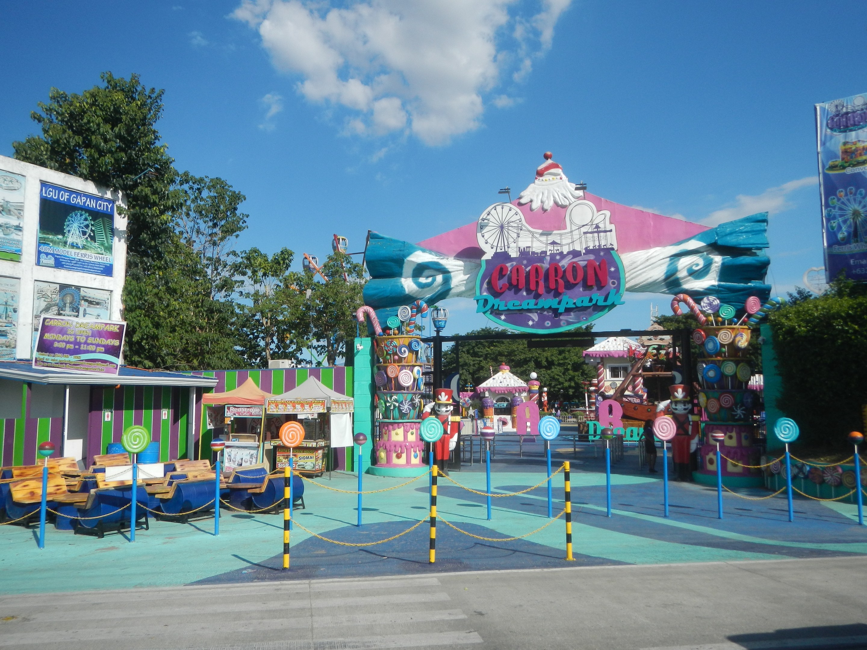 Kirmesfahrgeschäft am Parkeingang mit Straße, Zaun, Buden, Banner, Bäumen und bewölktem Himmel im Hintergrund.