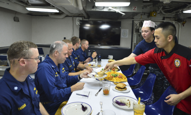 Eine Gruppe von Menschen teilt sich eine Mahlzeit an einem Tisch mit Tellern, Besteck und Getränken sowie einem Fernseher und Lampen im Hintergrund.