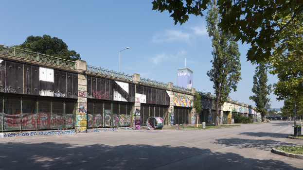 Leere Straße mit Graffiti an einer Gebäudewand, umgeben von Gebäuden, Straßenlaternen, Bäumen, Pflanzen, Gras und einem bewölkten Himmel.