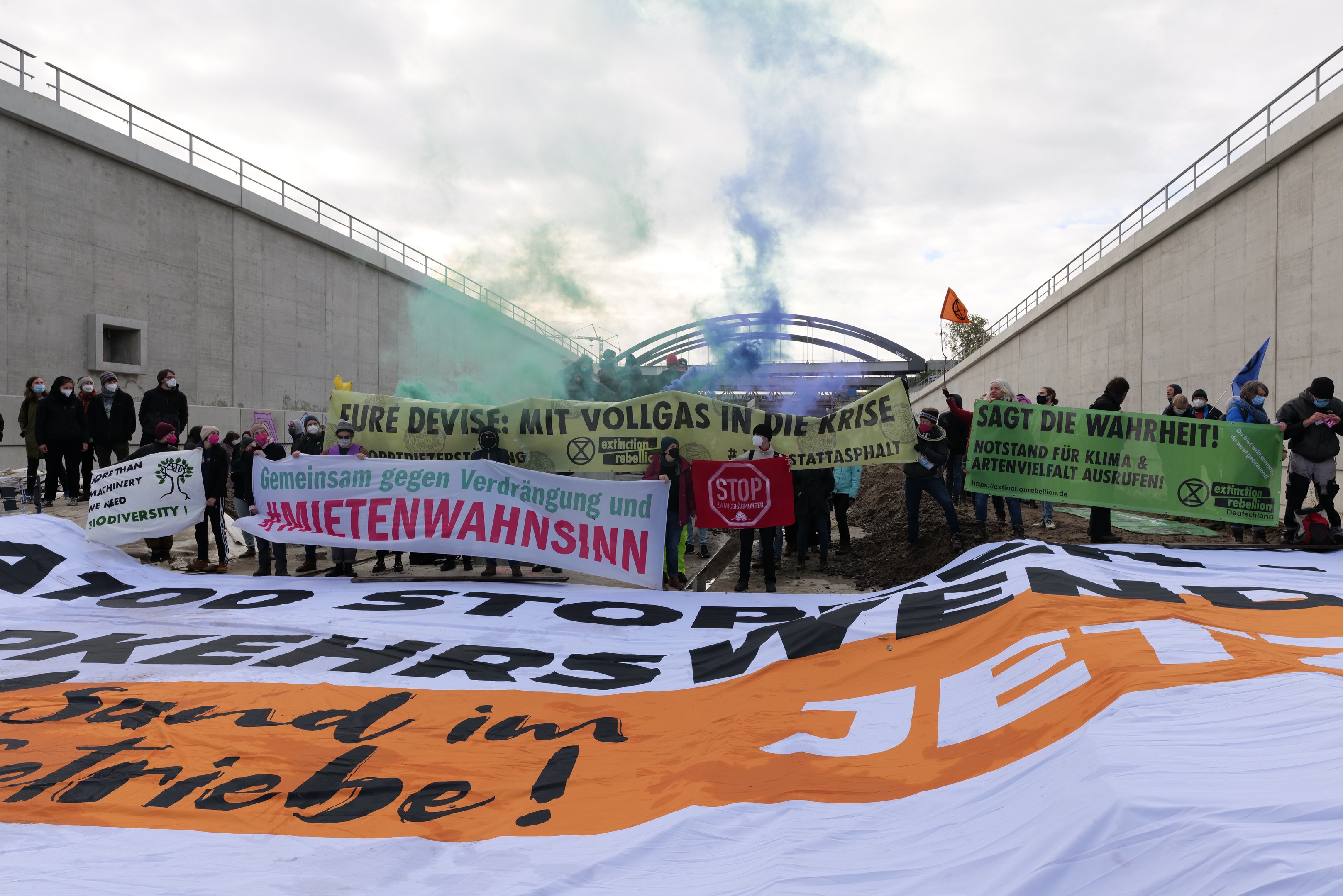 Menschen mit Schildern und Flaggen bei einer Klimaprotest in Berlin, mit einem großen Banner, auf dem "Klima-Protest in Berlin" steht und Rauch im Hintergrund zu sehen ist.
