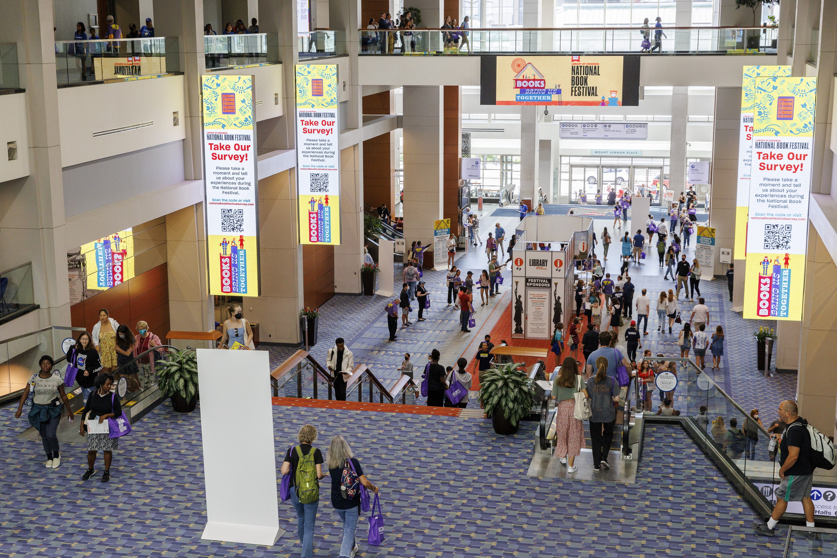 Eine große Menschenmenge, die durch ein großes Gebäude mit Bannern, Topfpflanzen, Rolltreppen, Geländern, Säulen, bedruckten Tafeln, Glastüren und anderen Gegenständen geht, wobei einige Taschen tragen, beim Nationalen Buchfestival.