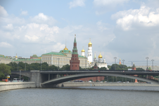 Eine Brücke mit fahrenden Fahrzeugen über einen Gewässser, flankiert von Laternenpfählen, mit Bäumen und Gebäuden im Hintergrund und dem Moskauer Kreml unter einem bewölkten Himmel.