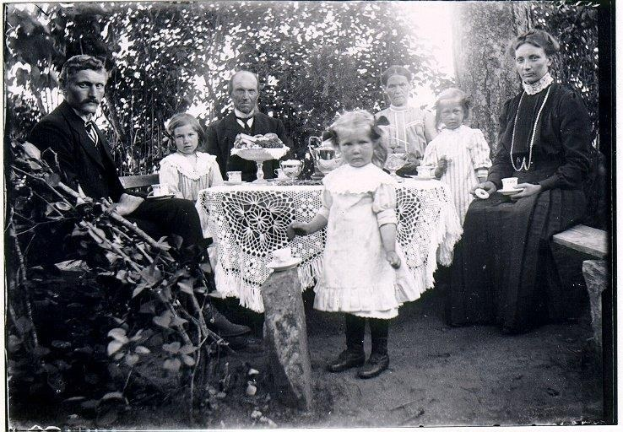 Schwarz-weiß-Foto einer Familie, die sich um einen Tisch in einem Garten versammelt hat, mit Tassen, Untertassen und anderen Gegenständen auf dem Tisch, umgeben von Bäumen und Pflanzen.