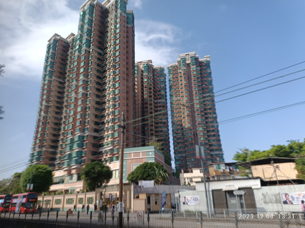 Eine belebte Stadtstraße in Mumbai mit hohen Wohngebäuden, Fahrzeugen, einem Zaun, Strommasten, Schildern, Bäumen und Passanten unter einem bewölkten Himmel.