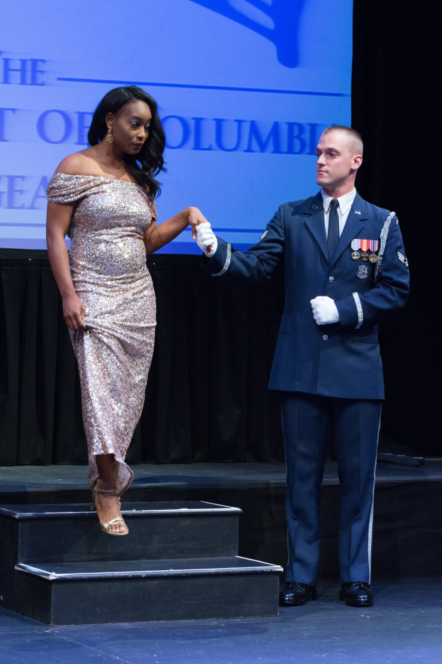 Zwei Personen auf einer Bühne, die Hände haltend, der Mann im Anzug und die Frau in Schuhen, mit einem Bildschirm und einem Vorhang im Hintergrund.