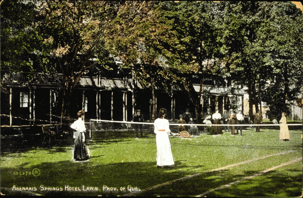 Black-and-white photo of a group of people playing tennis on the grass in front of the Abenakis Springs Hotel lawn in Provo, Quebec, with a net, chairs, trees, and a building in the background.