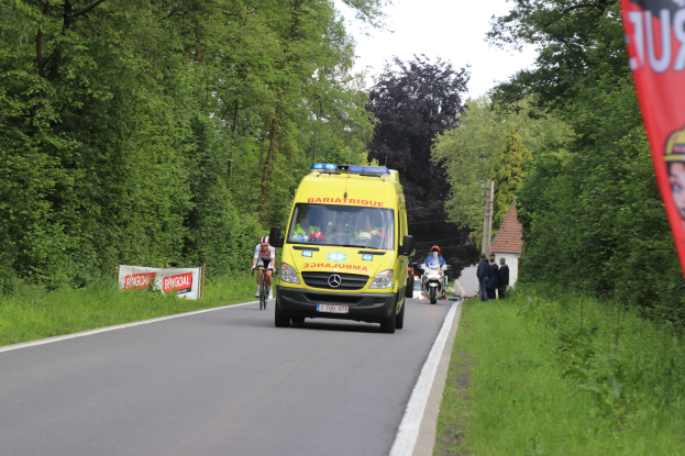 Ambulance driving down a road with cyclists alongside, grass and trees on either side, houses, poles, and a clear blue sky in the background.