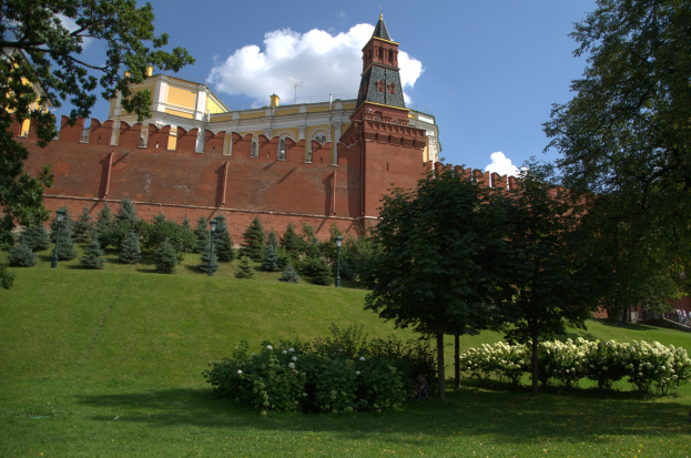 Roter Platz in Moskau, Russland, mit dem Kreml im Hintergrund, mit Menschen, Bäumen, Pflanzen mit Blumen, Straßenmasten und einem bewölkten Himmel.