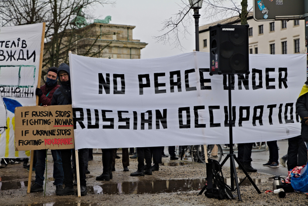 Eine Gruppe von Menschen mit einem Transparent, auf dem "Kein Frieden unter russischer Besetzung" steht, mit einem Redner, Taschen, einem Schild, einem Laternenpfahl, Bäumen, einer Statue, Gebäuden und einem bewölkten Himmel im Hintergrund.