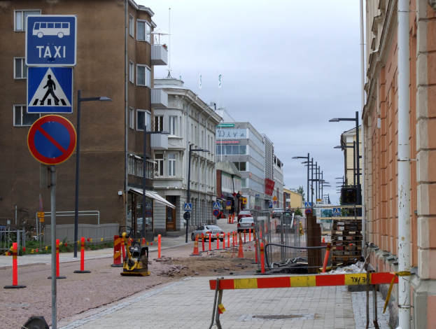 Stadtstraße mit Gebäuden, Straßenlaternen, Schildern, Verkehrskegeln, Fahrzeugen, Absperrpoller, Bäumen, einer Baustelle mit Verkehrsschildern und einem bewölkten Himmel.