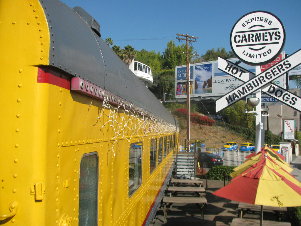 Ein mobiles Kantinenfahrzeug ist in der Nähe von Bänken und Treppen geparkt, mit Schirmen, Pflanzen und einem Geländer in der Nähe. Fahrzeuge, ein Pfahl, Bretter, Absperrungen, ein Gebäude, Bäume und verschiedene Objekte sind ebenfalls zu sehen, unter einem klaren Himmel.