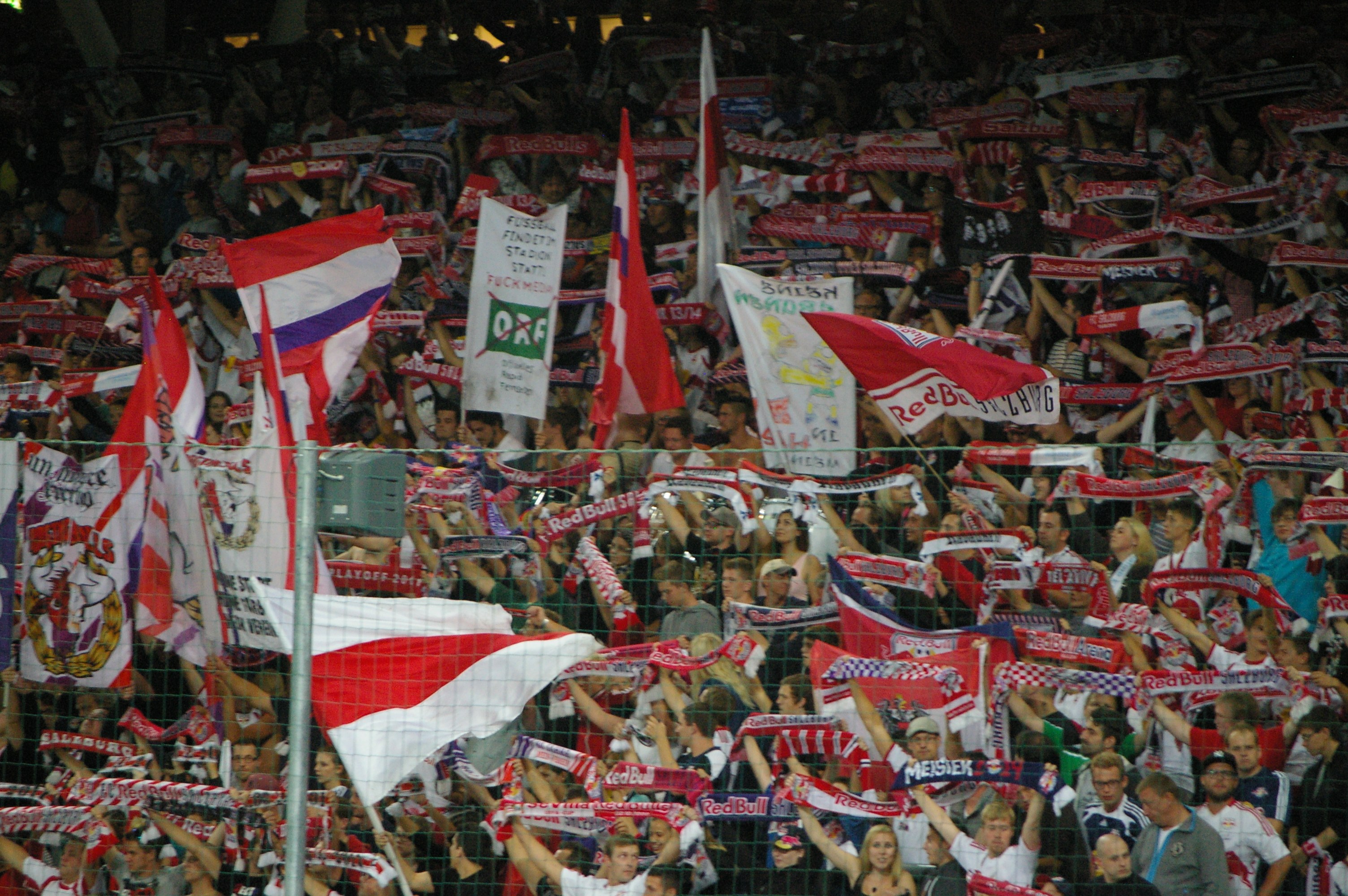 Große Menschenmenge in einem Stadion mit Menschen, die Fahnen und Banner halten, hinter einem Maschendrahtzaun stehend, mit Polen und Lichtern im Hintergrund.