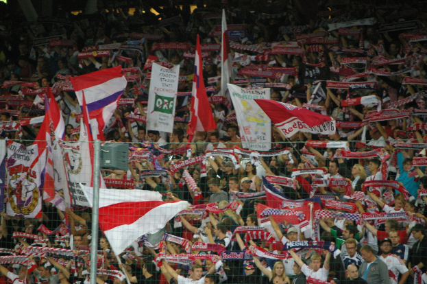 Große Menschenmenge in einem Stadion mit Menschen, die Fahnen und Banner halten, hinter einem Maschendrahtzaun stehend, mit Polen und Lichtern im Hintergrund.