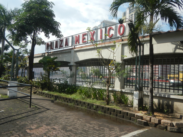 Der Eingang zum Plaza Mexico in Mexiko-Stadt mit einem Namensschild an der Wand, einem Metallzaun, Bäumen, Pflanzen, Gras, einem Weg, Pfählen, einer Straßenlaterne, Fahrzeugen auf der Straße, Gebäuden und einem bewölkten Himmel.