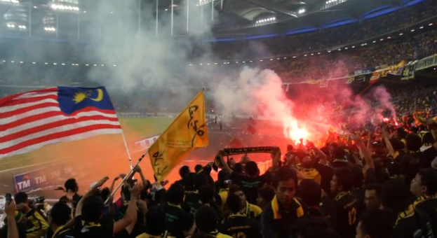 Eine große Menschenmenge in einem Stadion mit brennenden Fackeln in der Luft, einige halten Fahnen und eine Person hat eine Kamera, mit Rauch und Schildern im Hintergrund, die 'Malaysia vs Malaysia Live Streaming' lesen.